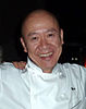 Masa Takayama, a bald-headed smiling middle-aged man wearing a chef's white double-breasted jacket with the name Masa on it