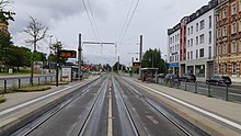 Straße. Mitte: zwei Straßenbahngleise und überdachte Haltestellen auf beiden Seiten. Rechts: drei Autospuren. Links: mindestens eine Autospur.