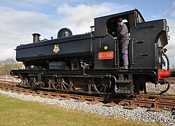 A pannier tank locomotive is standing on a siding. The locomotive is mainly painted black. On the side of the pannier tank is a crest consisting of a red and white spoked wheel. Across the wheel is a black and white label with the words "BRITISH RAILWAYS". A yellow lion stands on the label, above the wheel. On the side of the cab is a red number plate with the edge and number in brass. The rear buffer beam is red.