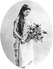 Photographie noir et blanc d'une jeune femme en robe, tenant un bouquet de fleur dans les mains et le regardant.