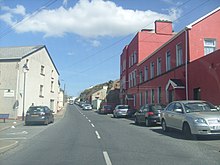 A road between two buildings, one red the other pebbledash, with cars parked on either side.
