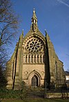 The west end of a Gothic Revival church with a small doorway, arcading, a large and elaborate rose window, and a narrow bellcote