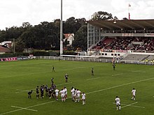 Deux équipes de rugby se font face-à-face avant de former une mêlée. En arrière-plan, la tribune d'un stade.