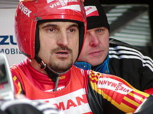 Georg Hackl in a helmet and luge gear, prepares for a luge run and confers with another man on his right.