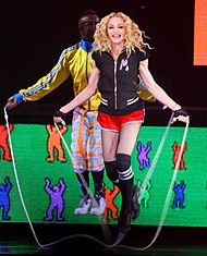 Picture of a blonde woman jumping rope. She's wearing a black vest, red shorts, and black knee-high socks. Behind her, a black male wearing a yellow-and-blue sweatshirt, and blue-and-orange pants, also jumps rope. The backdrop behind them is green and depicts different colored human-like drawings
