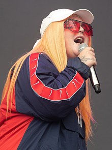 Tones and I is singing into a hand-held microphone with her eyes partly closed. The back of her right hand has an obscured image or a tattoo. She wears a white cap and rose-coloured glasses. Her hair is strawberry blonde and hangs down past her shoulders. She wears a red and blue jacket.