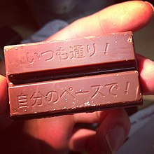 An unwrapped two-bar Japanese Kit Kat with text stamped into each bar. The text reads, "いつも通り!自分のペースで!"