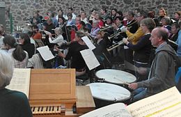 Baroque orchestra with prominent timpani (kettledrums) and trumpets surrounded by a choir in a rehearsal