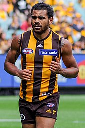 A male athlete with dark hair wearing a sleeveless jersey and shorts runs on the grass surface of the playing arena.