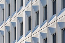 Des rangées de fenêtres toutes pareilles sur un mur blanc forment une géométrie de gris.