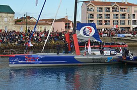 Vu de profil, le bateau bleu et gris descend le chenal. Shiraishi est près du mât en vêtement noir d'arts martiaux. Il y a foule sur les quais et aux fenêtres.