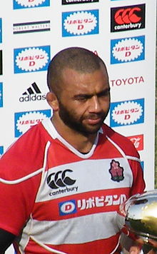 Le joueur, le maillot rouge et blanc du Japon sur les épaules, tient un trophée dans les mains devant un mur publicitaire.