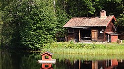 Ett brunt envåningshus med dubbeldörrar som står öppna. Bakom byggnaden finns träd och framför den ligger en damm.