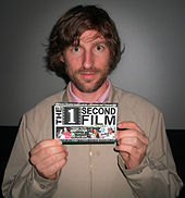 A man in a pink dress shirt and tan jacket holding up a producer credit for "The 1 Second Film" (as indicated by the text).
