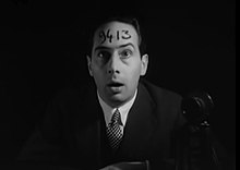 A man wearing a suit jacket and tie stares forward with a shocked and confused facial expression, with the numbers "9413" written across his forehead.