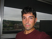 Dark-haired man with unkempt hair. He is wearing a red shirt. A silver chain around his neck and an earring in his left ear can be seen.