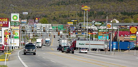 Route semi-urbaine à deux fois deux voies, surmontée de nombreux panneaux routiers et feux et entourée de publicité.