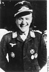 A smiling man wearing a peaked cap, military uniform with an Iron Cross displayed at the front of his uniform collar.