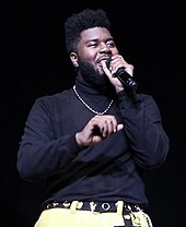 Khalid with a microphone in his left hand and his right hand raised midway