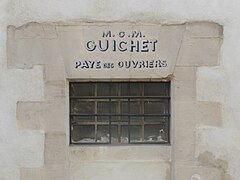 Fenêtre étroite percée dans un mur, sur lequel est inscrit « M.O.M. Guichet Paye des ouvriers ».