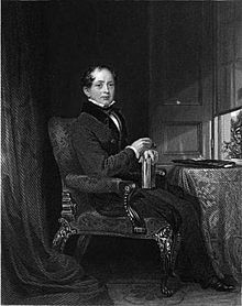 A black and white portrait of man wearing a black jacket and white shirt with a black bow tie. He is partly bald with short, wavy hair around the sides of his head. He is sitting on a chair with a book on his lap, his right hand on top of the book, and his left hand resting on top of his right. A table is to his right and a partially open window sits above and to the right of the table. A dark curtain is behind him.