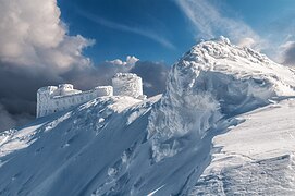 Bâtiment recouvert de neige sur une crête, ciel bleu et nuages.