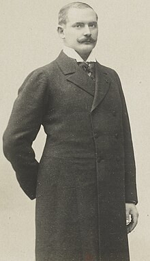 Black and white photo of a man with a thick moustache wearing a long black coat