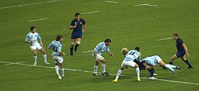 Photographie en couleurs. Sur un terrain, une équipe en bleu nuit affronte une équipe en maillot rayé de blanc et bleu ciel : un joueur bleu nuit est en train de se fait plaquer par deux joueurs de l'autre équipe.