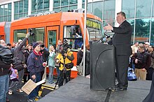 Mayor Greg Nickles speaking from a podium to a crowd of people, including several camera operators, with an orange streetcar in the background.
