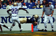 A player pursued by an opponent dives into the end zone in front of an official to score a touchdown.