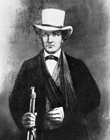 Portrait d'un homme portant une veste sombre avec un chapeau blanc. Son bras gauche est en écharpe et il tient un fusil dans sa main droite.