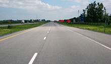 "A look down a rather level two-lane asphalt highway, with cars and trucks at some distance traveling in the same direction. Oncoming vehicles use the two-lane roadway some 3 or 4 metres to the left, separated by a grassy median. Orange signs and barrels to the right indicate construction work and farther to the right, a tall blue-and-white sign and flags welcome motorists to Quebec."