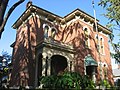 Red brick two-story house with a second-story balcony, stair leading up to the entrance coved by a striped awning, tall narrow windows, and small fence in front