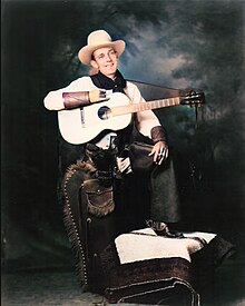 Studio picture of Jimmie Rodgers smiling and wearing cowboy attire: he dons a white Stetson hat, a short scarf on his neck, a two-tone western shirt, leather wrist cuffs, leather chaps, a revolver on the right side of his hips, cowboy boots, and spurs. A guitar hangs on his right shoulder from a strap as he holds it horizontally with his right arm. His left arm rests on his raised left knee and he holds a cigarette with his left hand. While his left foot rests on a stool covered with rugs his right leg stands straight