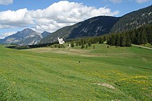 La photo couleur présente un alpage fleuri et vallonné au premier plan. En arrière plan, un monument honorant la Résistance dans le plateau des Glières se découpe face aux versants boisés et rocheux du fond de l'image.