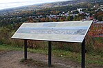 A long, wide photo rests upon three support columns on the face of a steep hill. A hilly landscape and river, with city housing, can be seen in the background.