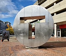 A large metal disk with "T" cut out from the middle, in front of a parking garage