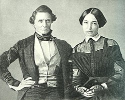 man on left with right hand on his hip; woman on right with both hands in her lap