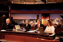 Four people seated at a news desk on a set