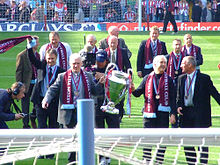 In the foreground is two men holding a large cup, they have claret scarves and a medal around their necks. Around them are ten old players in suits with medals and scarves around their necks