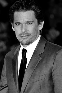 A caucasian male with dark slick hair, wearing a two-piece grey suit, with a white shirt and black tie.