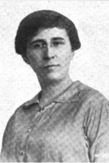 A photograph of a white woman with dark hair; she is wearing eyeglasses and button-down dress or blouse with an open collar