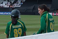 Two female in green cricket uniforms with yellow piping are talking. The left-hand female is wearing a helmet and has dark skin, 'ISMAIL' and '89' are visible in yellow writing on her back. The right-hand female has 'KAPP' and '06' visible on her back.