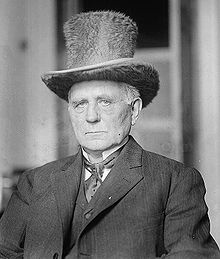 Clark dressed in a suit and tie and furry top-hat with an austere expression