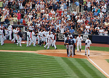 A man in a white baseball uniform with navy pinstripes and the number "2" on the back of his uniform runs towards home plate, while his teammates run to meet him to celebrate.