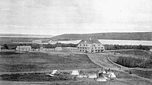 Exterior view of Qu'Appelle Indian Industrial School in Lebret, District of Assiniboia, c. 1885. Surrounding land and tents are visible in the foreground.