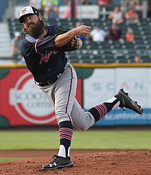 A man in a navy blue baseball jersey, gray pants, and a navy cap is seen at the end of his pitching motion having just pitched a ball from the mound.