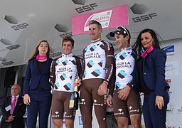 De trois-quarts, cinq personnes sourient. Les trois coureurs au centre portent des maillots de la même couleur.