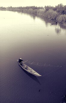 Photo d'un pêcheur dans sa barque vu dans. Il ramène un filet qui était immergé dans l'eau. Plus haut, un autre pêcheur déplace sa barque depuis la berge