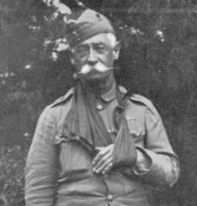 A black and white photo of a man with an impressive white mustache. His arm is in a sling and he is wearing a military cap.